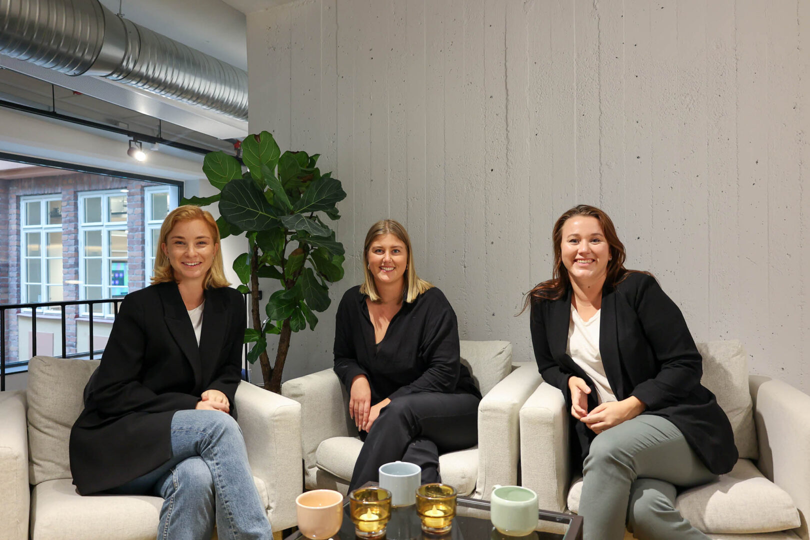 En Financial Controller, Compliance Officer och Risk Manager på ett tillväxtbolag sitter i varsin fåtölj i ett industriellt kontorslandskap och ler mot kameran, stolta över att ha säkerställt finansiell stabilitet och uppfyllt regulatoriska krav under en framgångsrik transformationsfas.