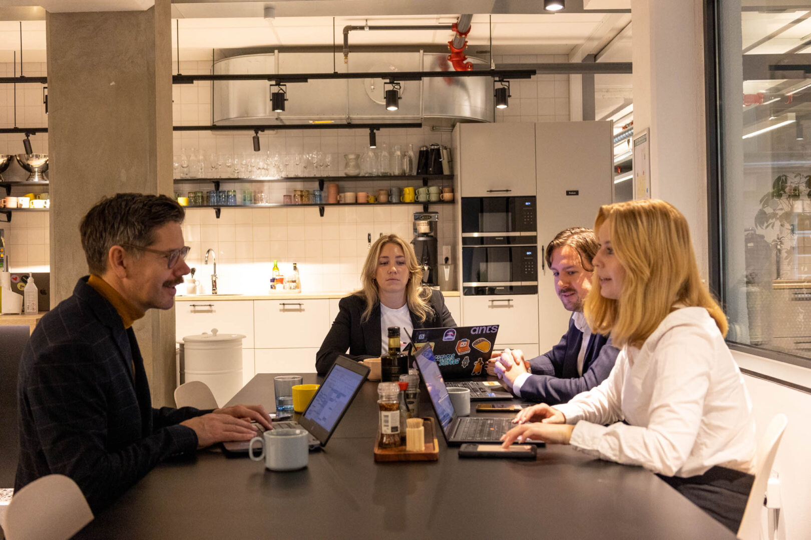 Fyra chefer, inkl. en CTO, COO, CDO och CISO sitter vid ett bord i ett kontorskök med sina laptops framför sig och diskuterar strategier för att optimera realtidsinfrastruktur och säkerställa datahantering i ett företag inom FinTech.
