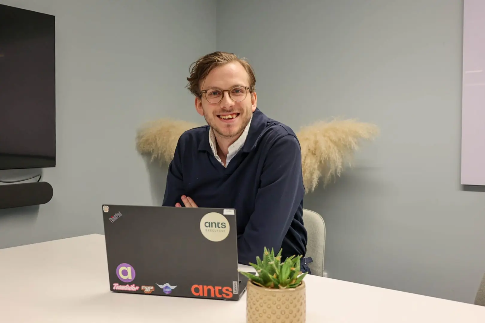 En Data Engineer sitter i ett ljusgrönt mötesrum framför sin laptop och ler in i kameran, glad över att bidra med sina expertkunskaper för att optimera digitala lösningar och förbättra produktionssystem.